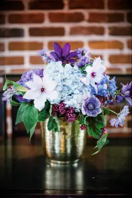 small wedding centerpiece in gold vase with purple clematis, anemones and blue hydrangeas