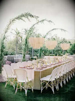modern outdoor wedding reception long banquet table with white riviera chairs and fringe chandeliers hanging from greenery arches