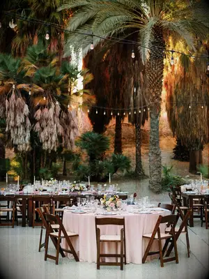 round banquet table with blush table cloth and dark wood folding chairs surrounded by palm trees