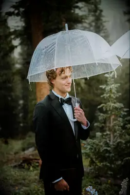 Groom in Classic Black Tuxedo Standing Under Clear Umbrella With Vow Book at Outdoor Ceremony