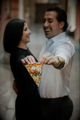 Couple holds out a slice of pizza while embracing. 