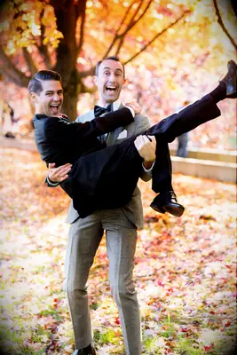 Two newly-engaged men smile in a fun pose among the autumn leaves