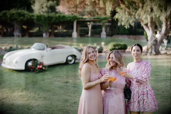 Three women raise a toast in front of a vintage car