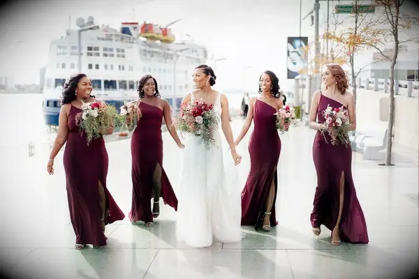 A bride walks with her bridesmaids by the water with a boat in the background.