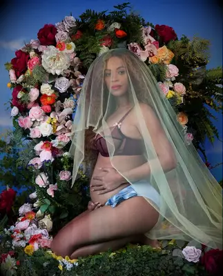 Beyonce poses for a maternity photo shoot with a lush floral background and a veil over her head. 
