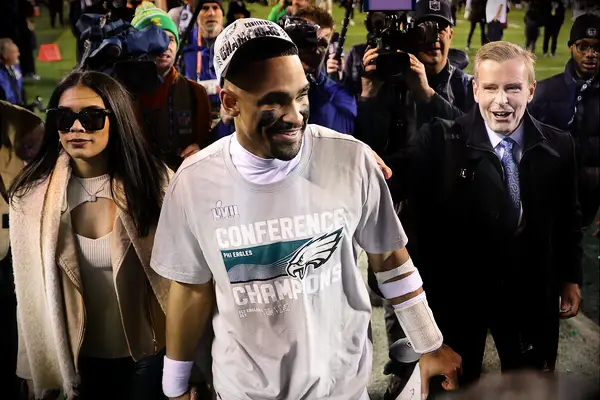 Jalen Hurts smiles and walks off the field hand in hand with a woman while reporters film. 