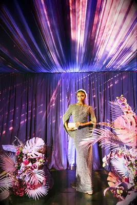 A bride poses for the camera in a stunning silver gown, standing among bright pink monsteras and ferns.