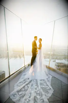 A happy couple stands on a glass promontory overlooking the big city, as the sun rises.