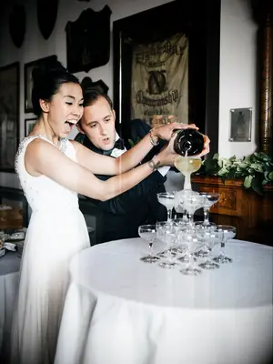 Bride and groom pop a bottle of champagne and poured it over a wedding champagne tower. 