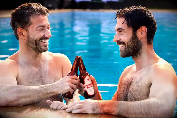 A gay couple enjoys some beers by a swimming pool