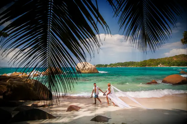 Newlyweds hold hands while walking down a tropical beach. 