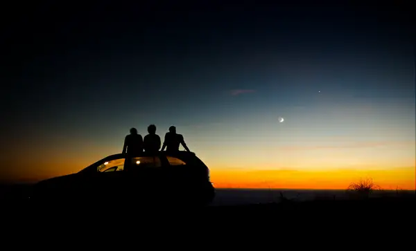 Three friends sit on the roof of their car and watch the sun set